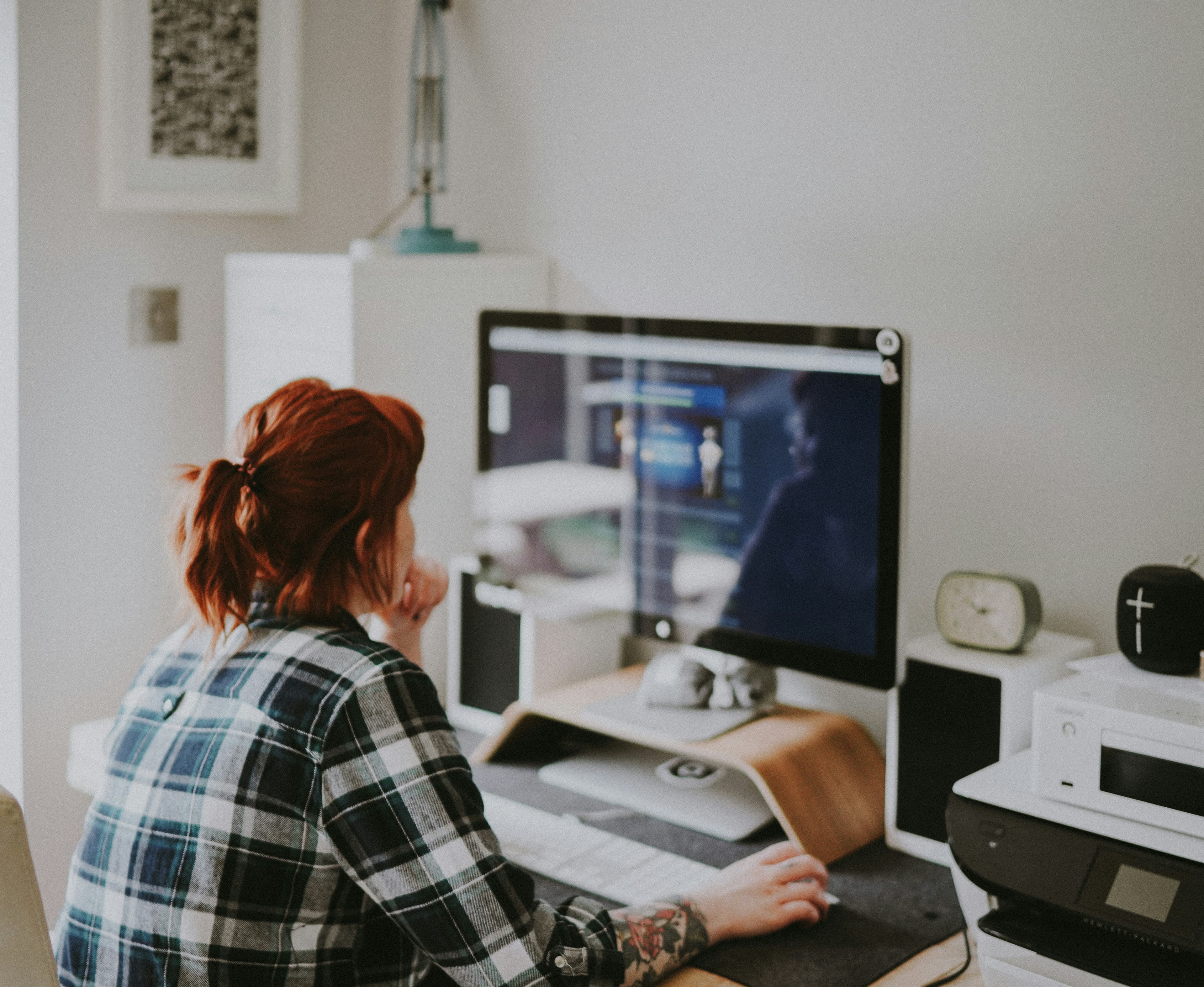 Person with red hair and a tattoo in a plaid shirt works at a computer.