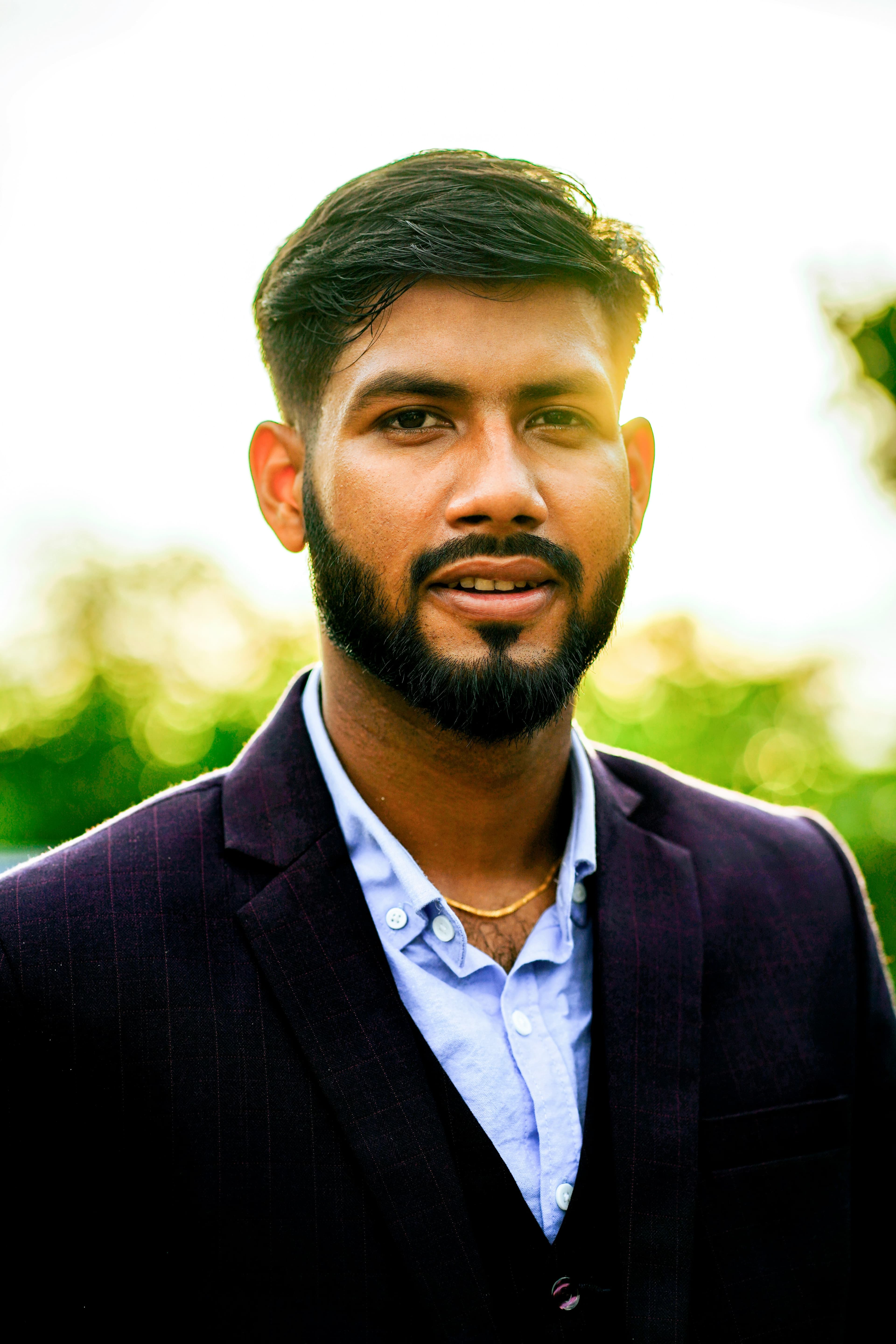 Close-up portrait of a bearded man in a purple blazer with a sunlit background.