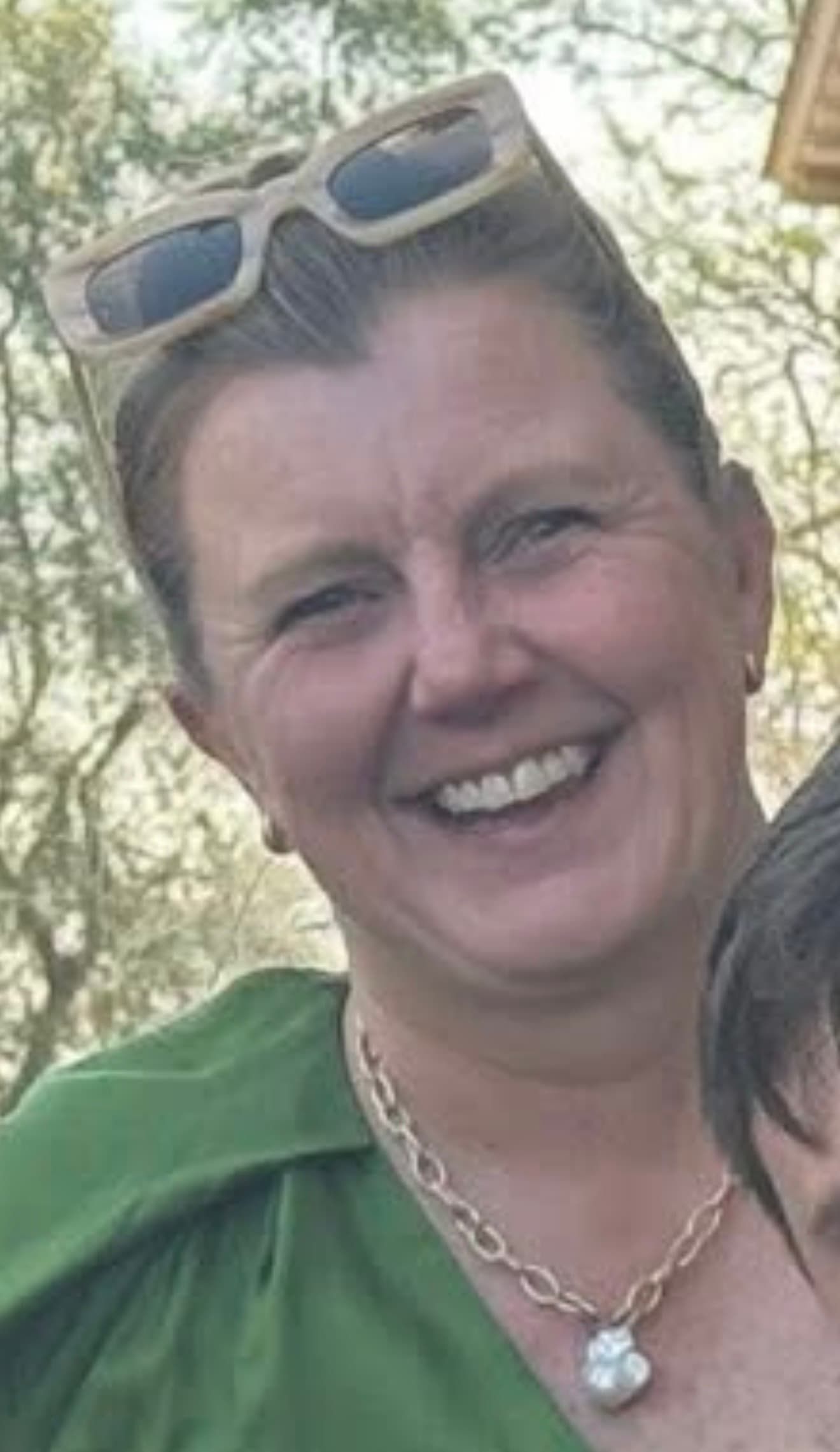 Smiling woman with sunglasses on her head, wearing a green top and pearl necklace.